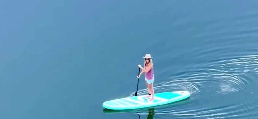 Person paddleboarding on calm lake, wearing hat.