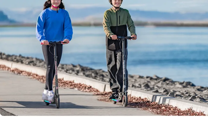 Two people riding scooters by a waterfront path.