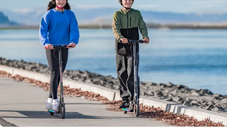 Two people riding scooters by a waterfront path.